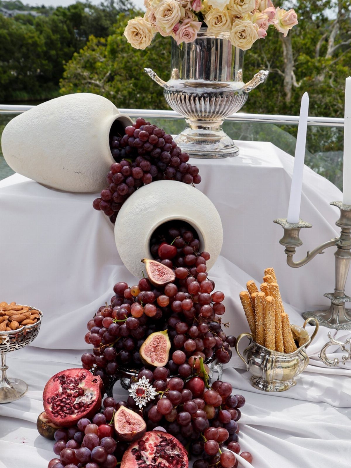 Festive tabletop display with spilling red grapes, figs, pomegranates, and silver serving pieces.