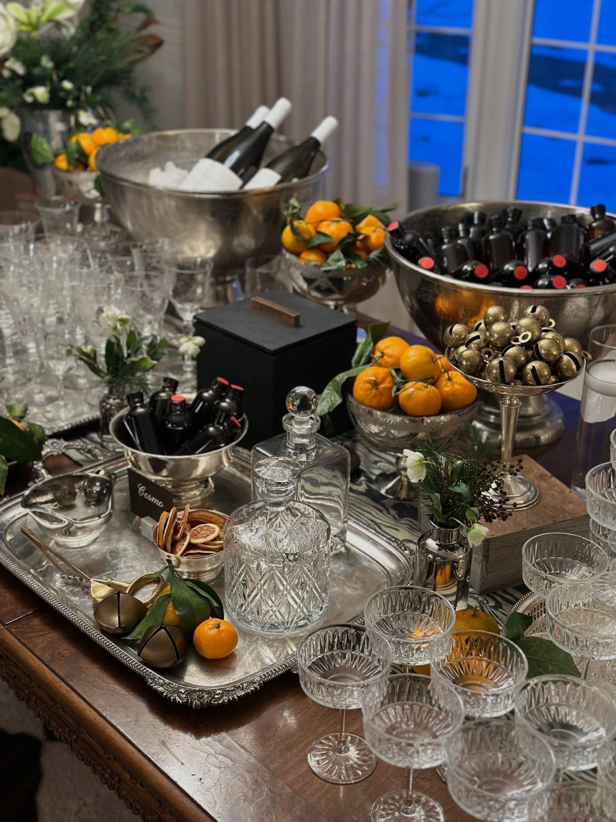 Drink station on a wooden table with silver bowls, crystal glassware, and oranges.