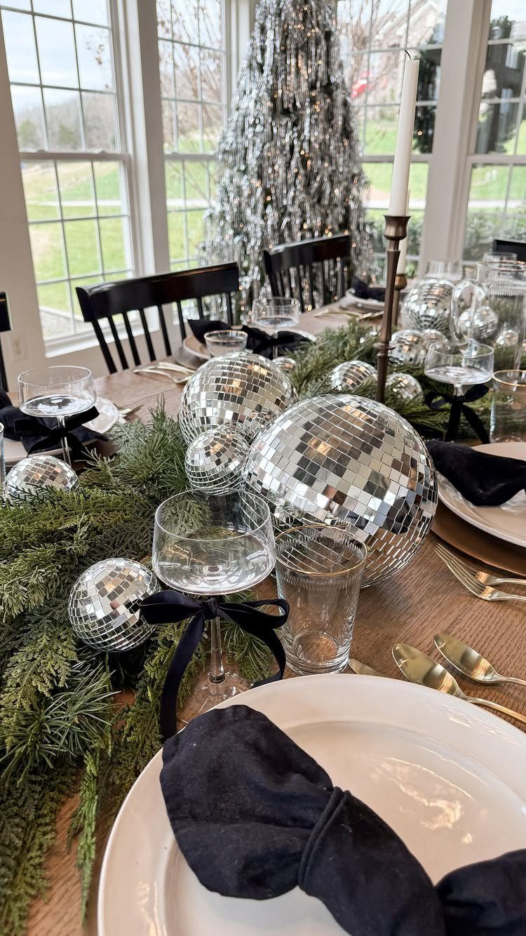 Dining table decorated with silver disco balls, greenery, and white plates for New Year's.
