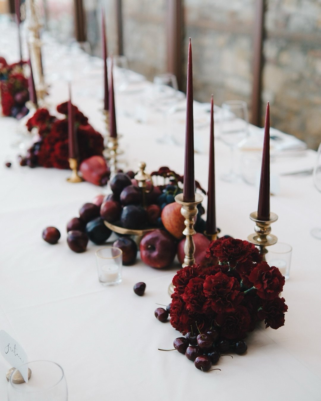Deep red flowers and tall taper candles arranged with dark fruits on white tablecloth