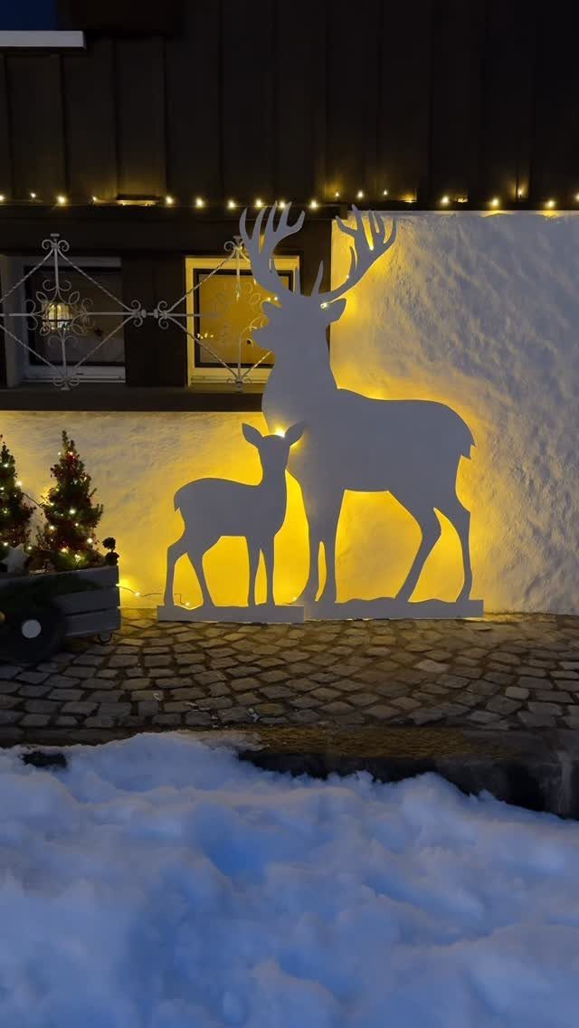 White reindeer silhouettes backlit with warm yellow light standing on a snowy cobblestone path.