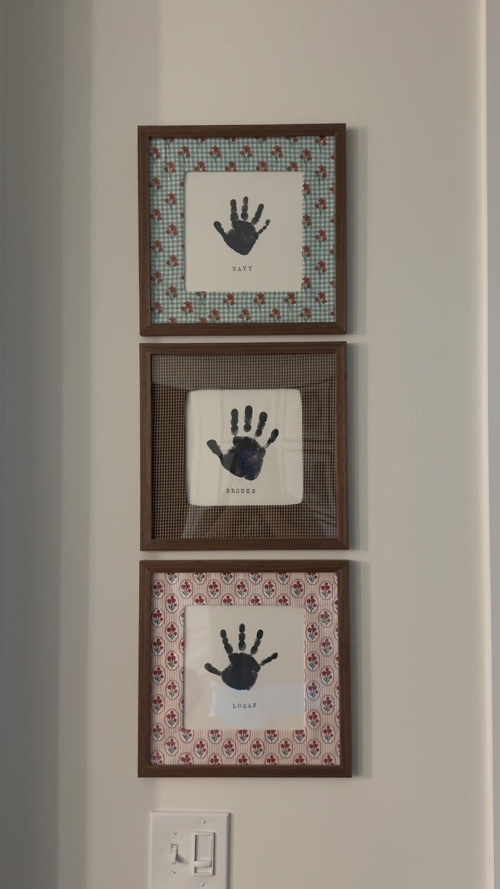 Three framed child handprints with names on a white wall as Mother's Day crafts.