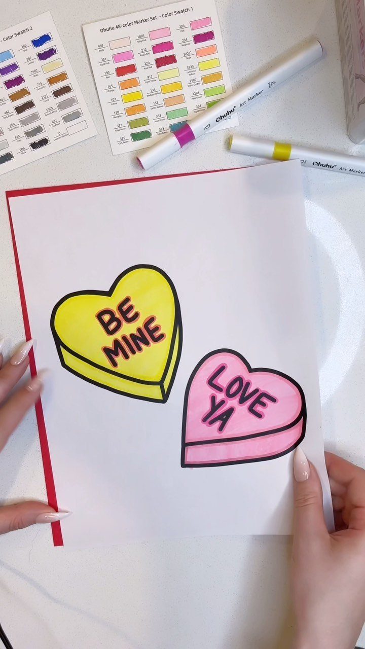 Two colored heart candies on a page held by hands with markers nearby.