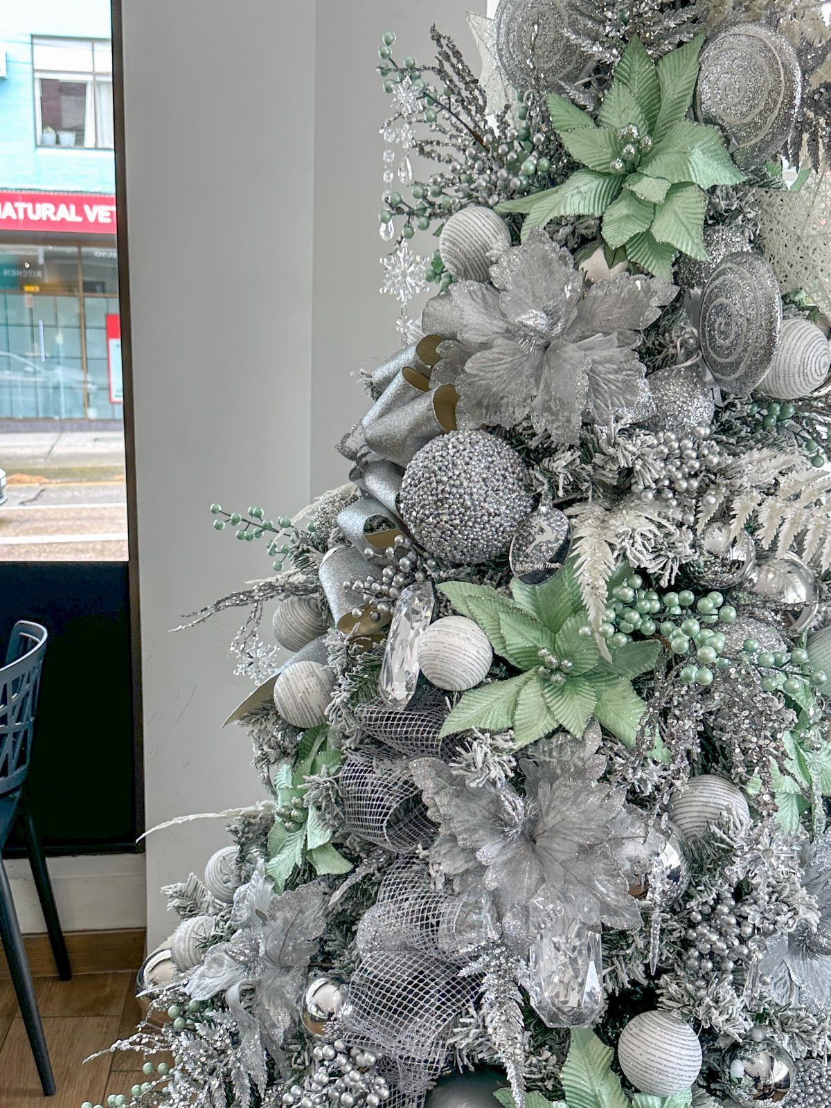 Decorated Christmas tree with silver ornaments, green floral accents, and silver mesh ribbon.