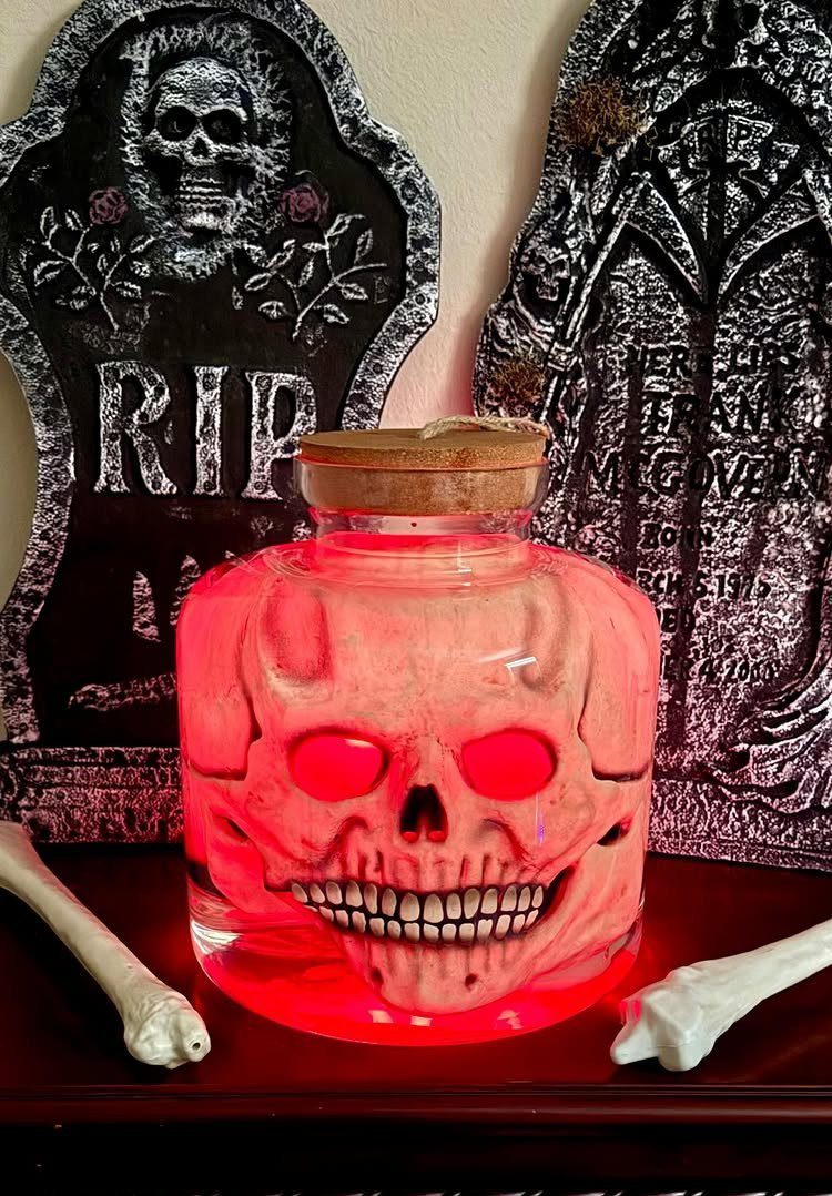 Red glowing skull inside a glass jar placed between two decorative tombstones.