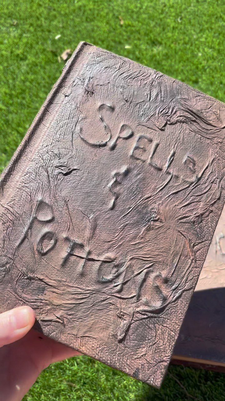 Brown textured spell book with embossed text held against a green grass background