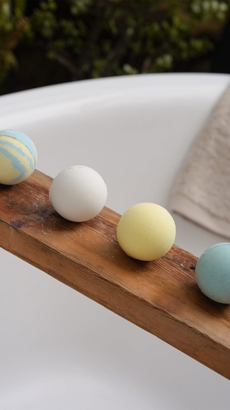 Four colorful spherical bath bombs resting on a wooden tray across a white bathtub.