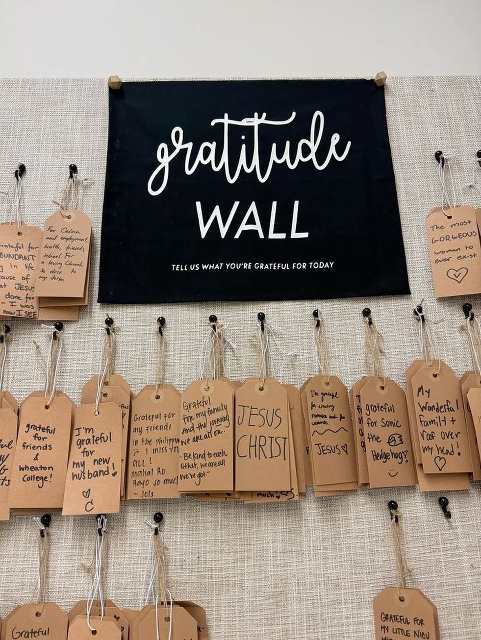 Gratitude wall with a black banner and handwritten brown paper tags on burlap.