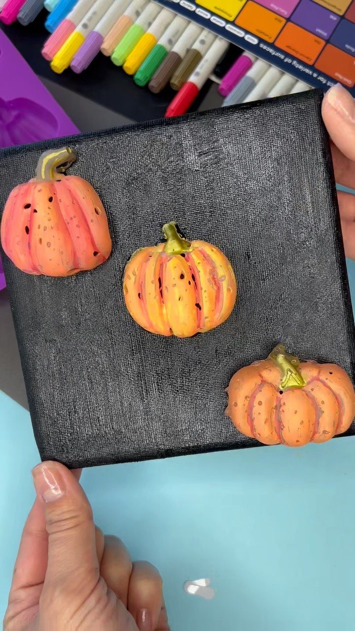 Three small painted orange pumpkins attached to a black square canvas held by hands.
