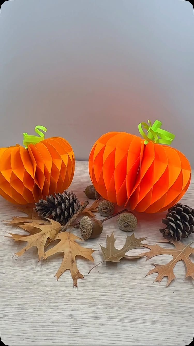 Two orange honeycomb paper pumpkins surrounded by autumn leaves, pinecones, and acorns.