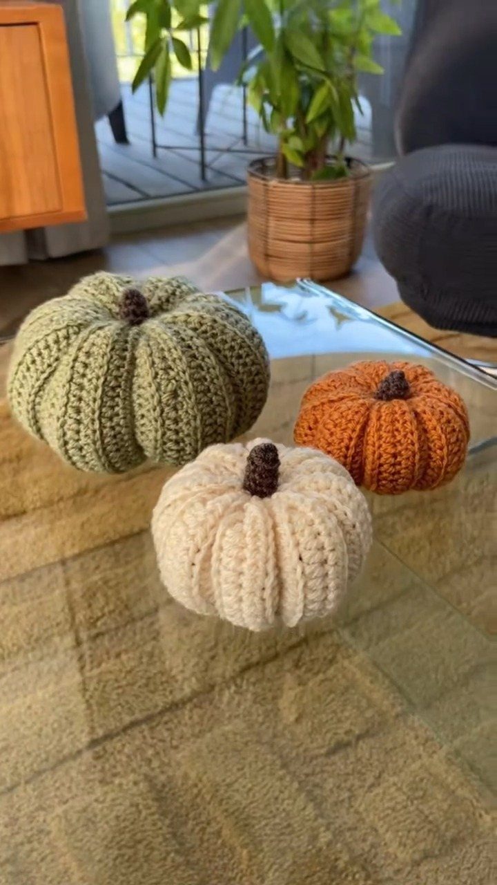 Three crocheted pumpkins in green, orange, and cream on a glass table.