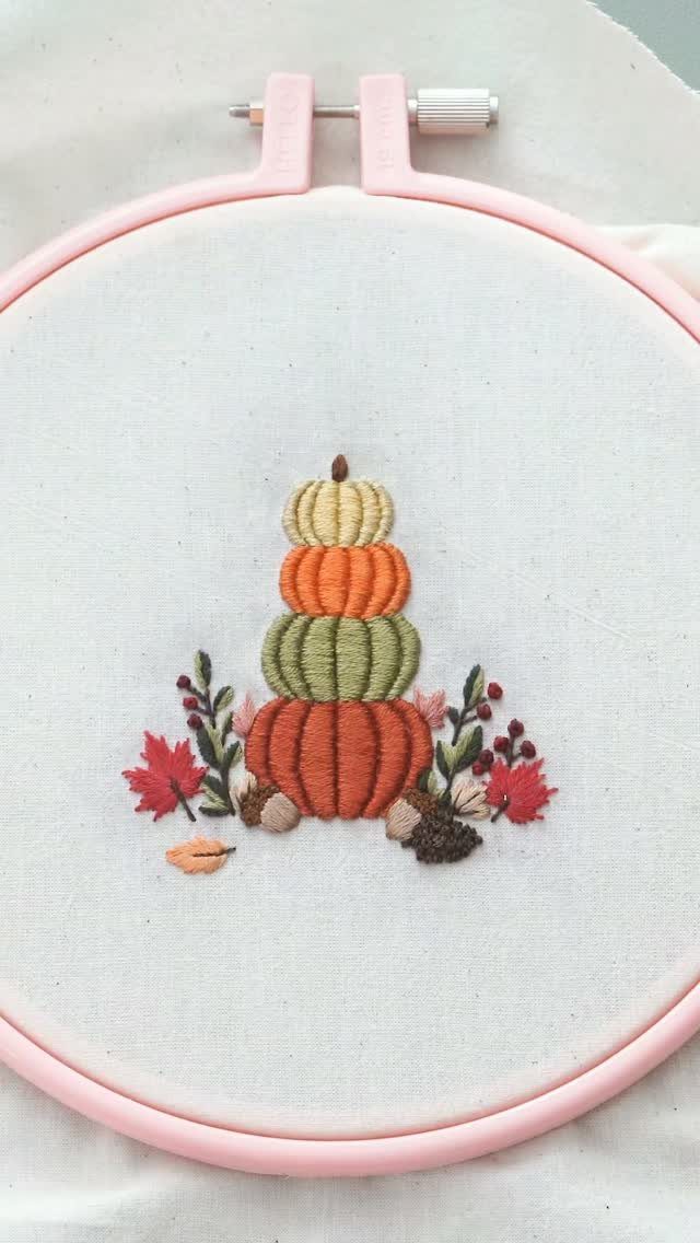 Stacked colorful pumpkins and autumn leaves embroidered on white fabric in a pink hoop
