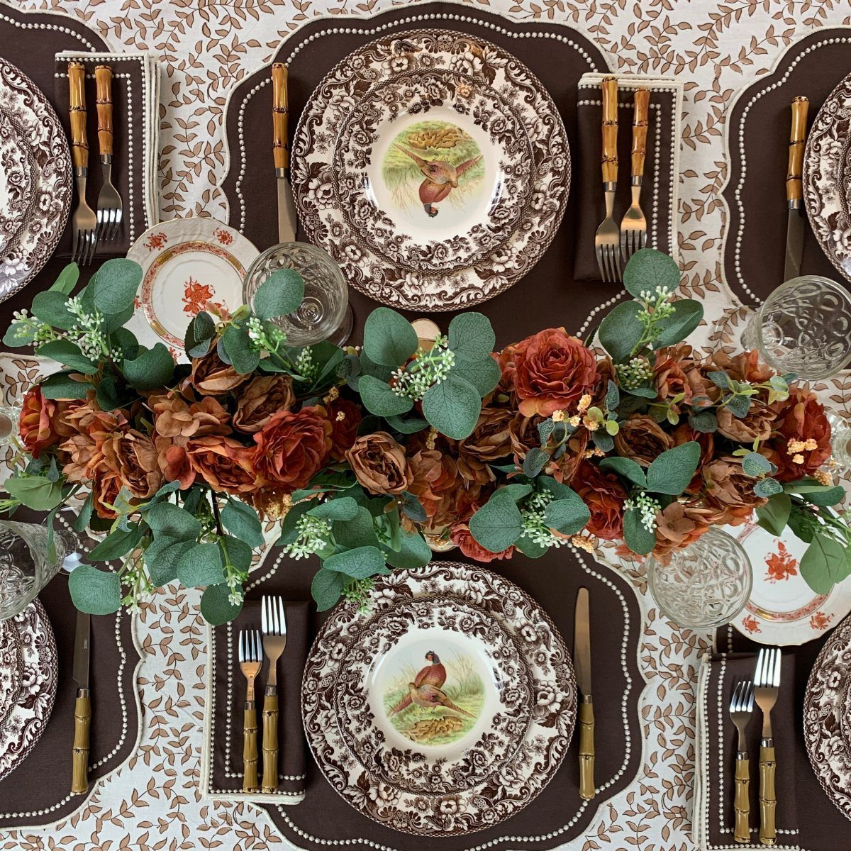 Set dinner table with orange rose centerpiece and brown patterned plates on floral cloth
