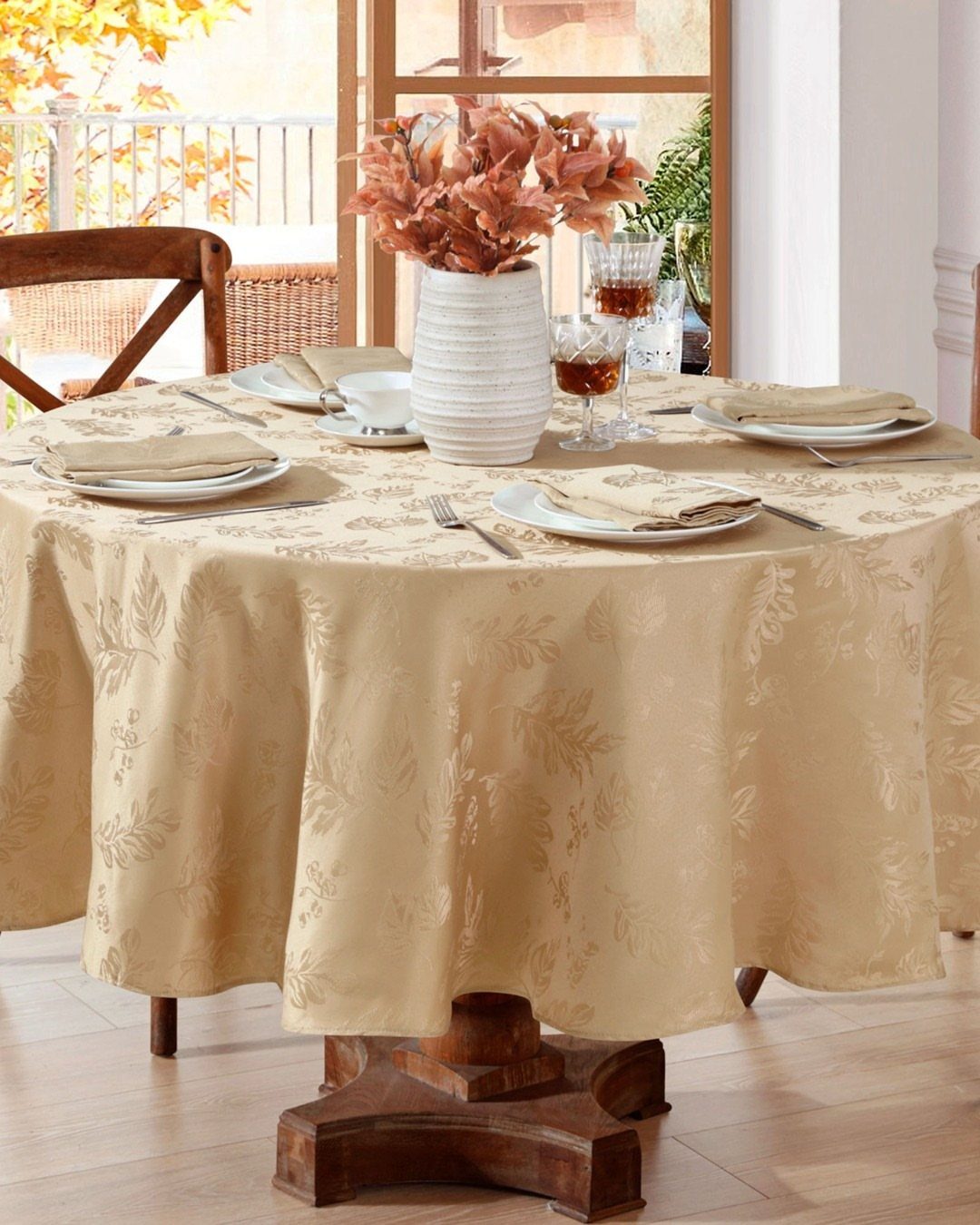Beige patterned tablecloth on a round wooden table with autumn centerpiece and place settings.