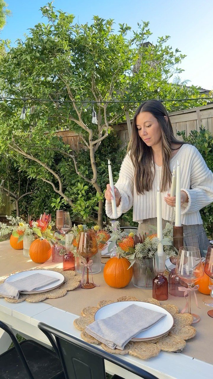 Woman arranging candles on a Thanksgiving table with pumpkins and amber glassware outdoors.