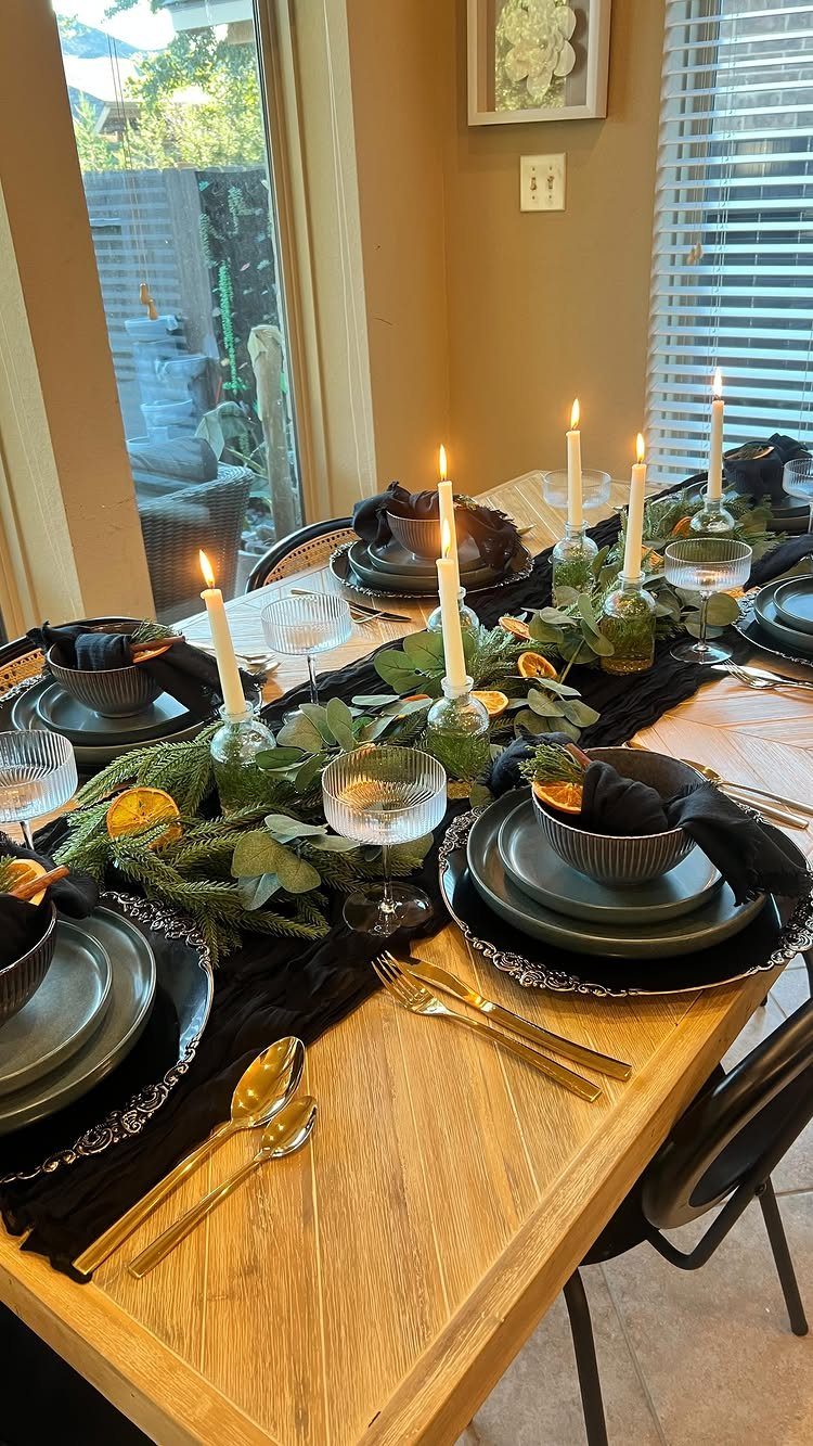 Wooden dining table with black runner, gold cutlery, and greenery centerpiece with candles.