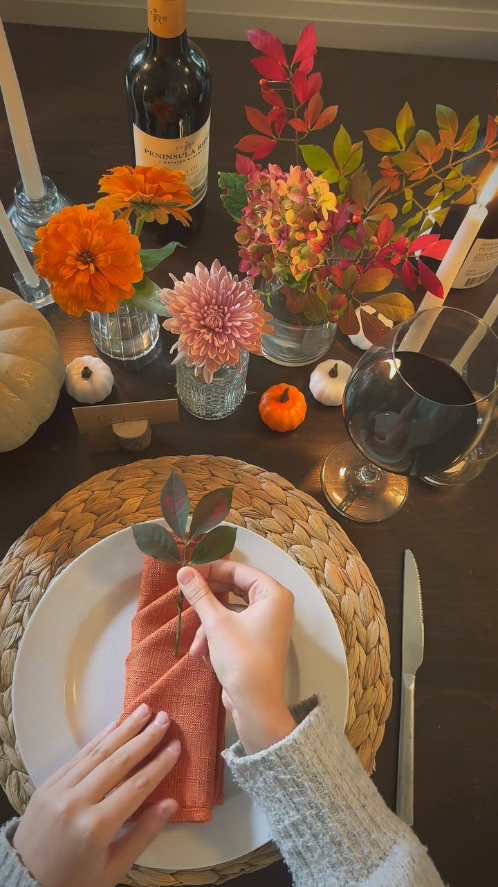 Hands placing a leaf sprig on an orange napkin at a Thanksgiving table.