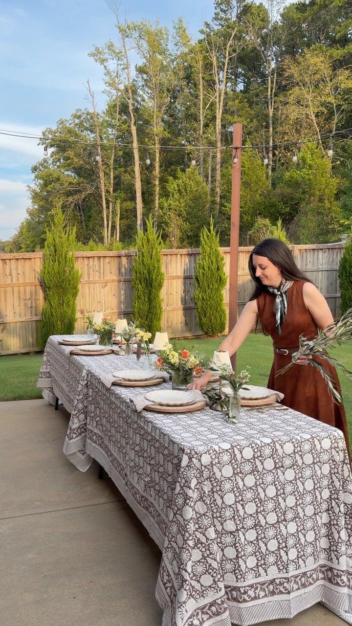 Woman arranging a decorated outdoor dining table with floral centerpieces and patterned linens.