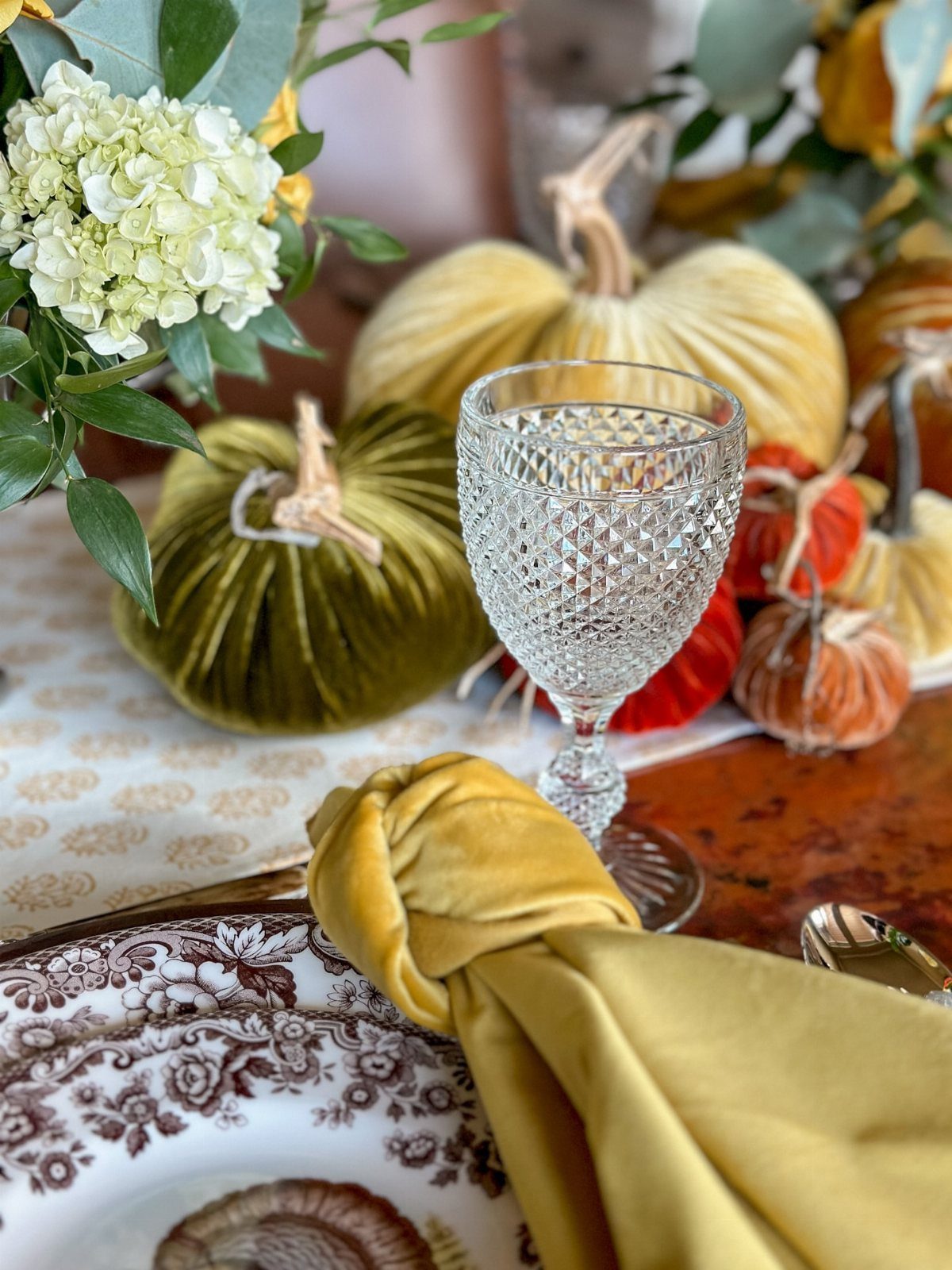 Patterned plate with gold velvet napkin, crystal glass, and velvet pumpkins on table.