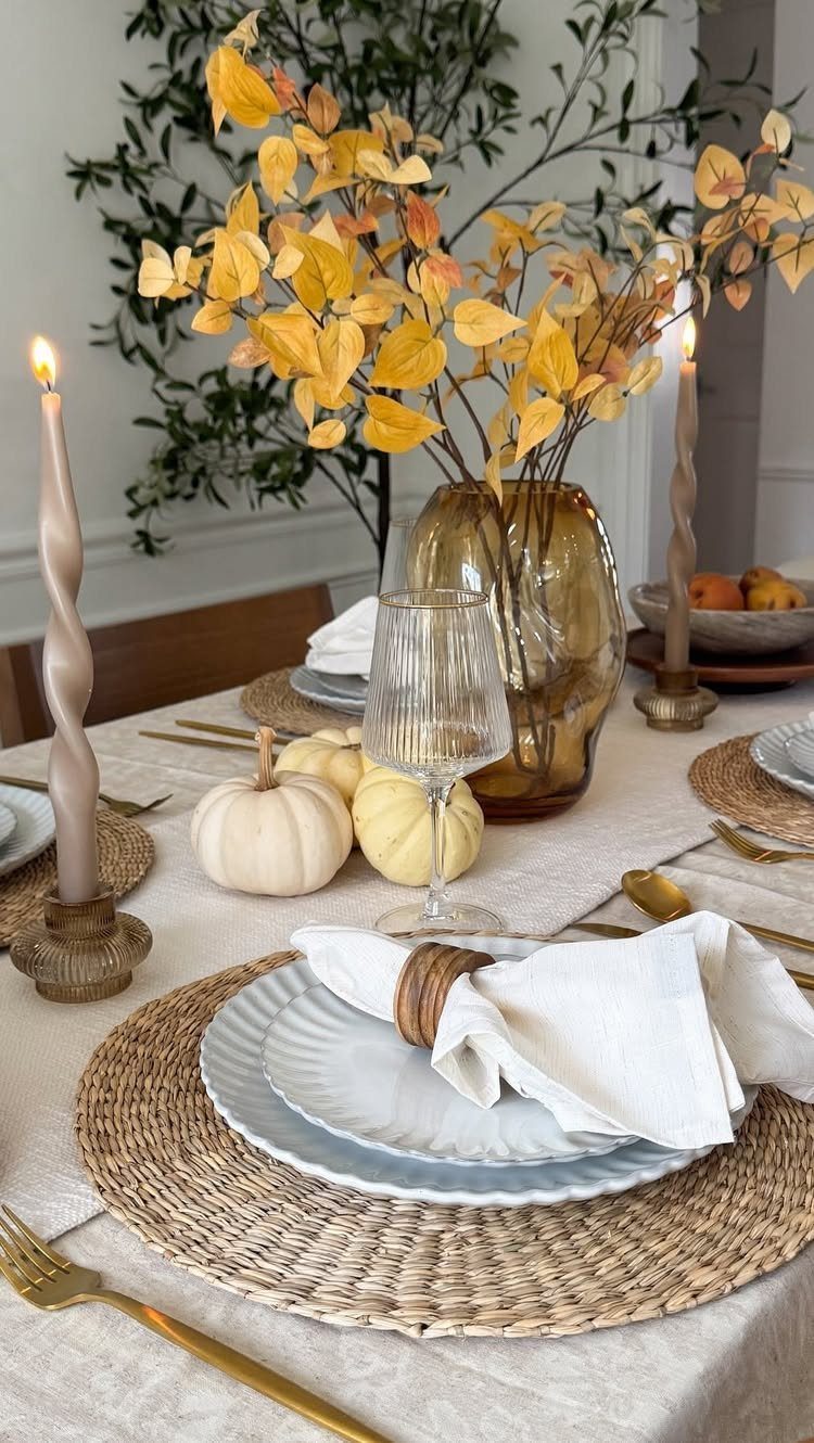 Thanksgiving table setting with white plates, woven chargers, gold cutlery, and autumn leaves.