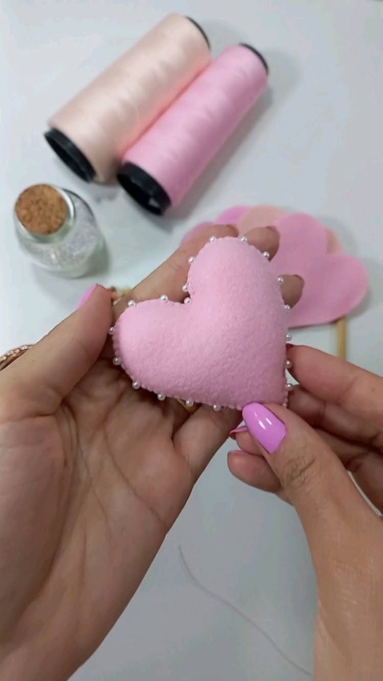 Hands holding a pink felt heart decorated with white pearls and sewing supplies.