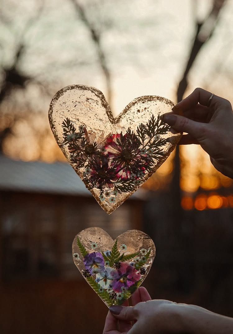 Heart-shaped resin ornaments with pressed flowers held up against a sunset background.