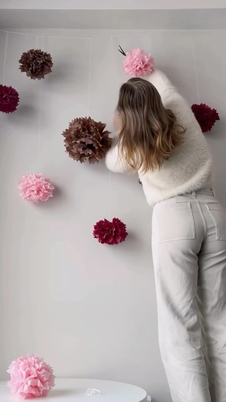 Woman hanging pink, red, and brown tissue paper pom-pom flowers on a wall