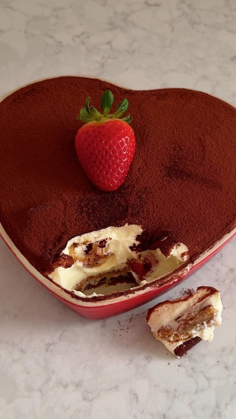 Heart-shaped red baking dish with tiramisu topped with a fresh strawberry on marble.