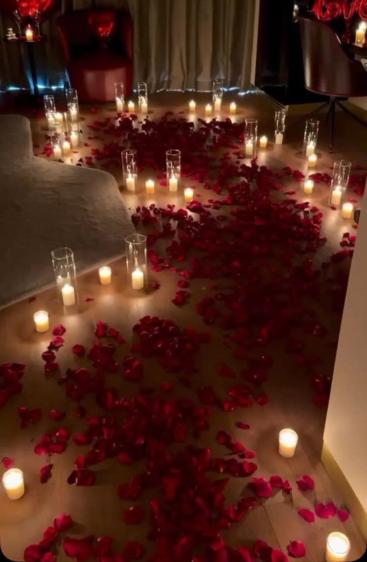 Red rose petals and glowing white candles scattered across a wooden floor