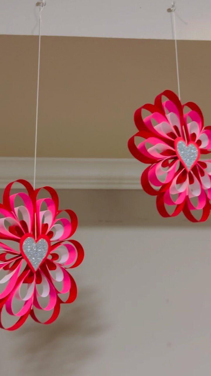 Paper flower decorations with silver hearts hanging from a ceiling by white strings.
