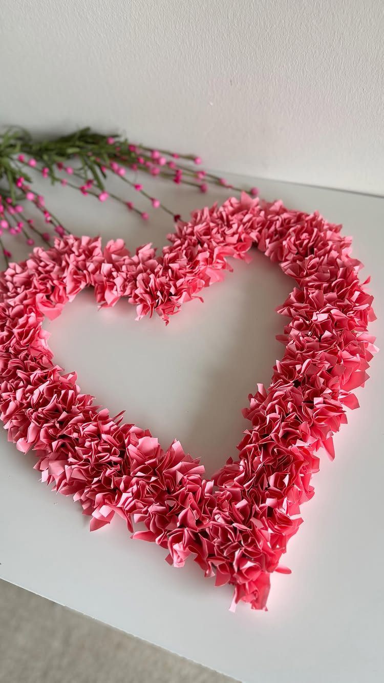 Pink ruffled paper heart wreath resting on a white surface with floral accents.