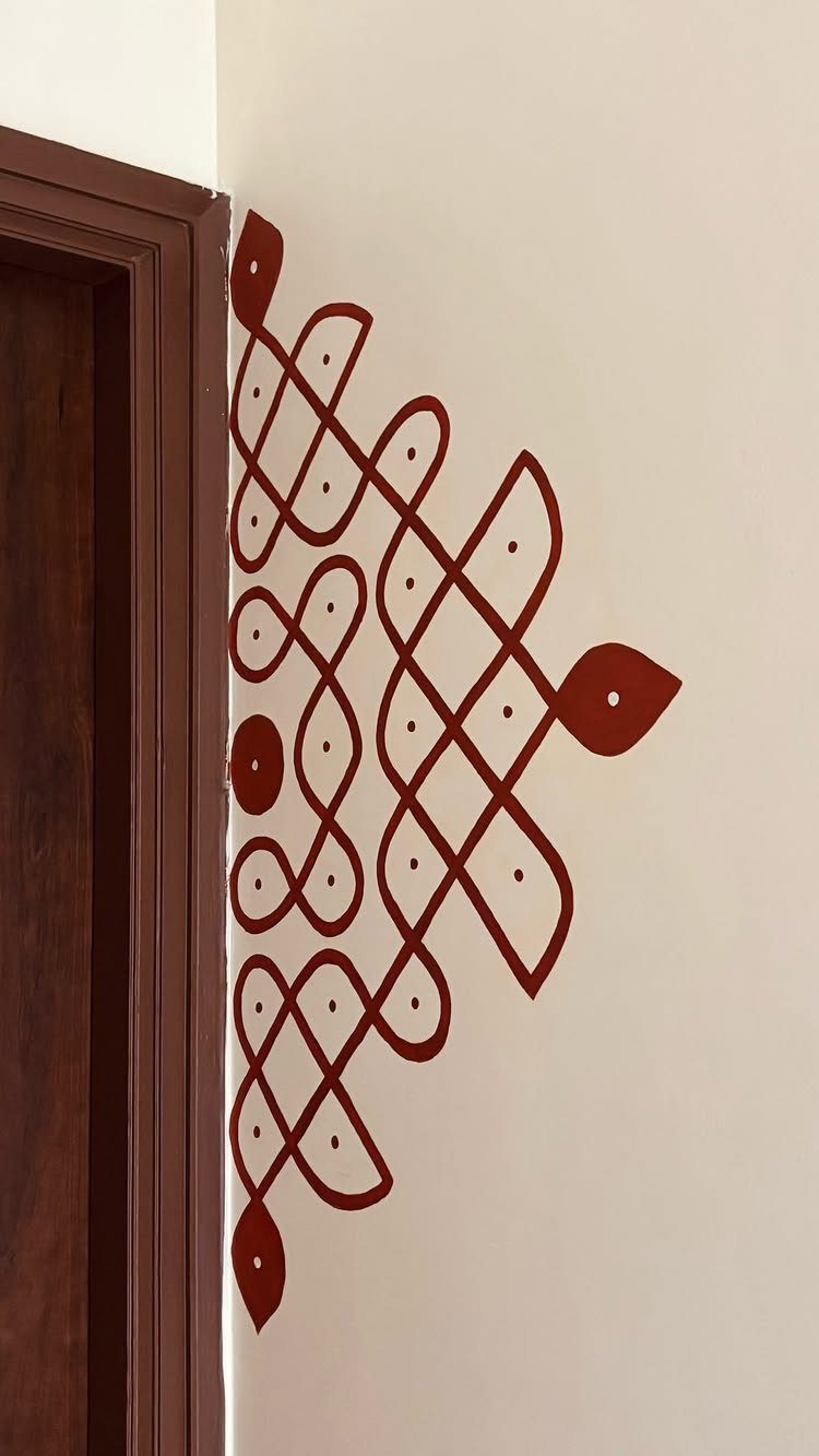 Reddish-brown traditional rangoli pattern painted on a white wall beside a wooden door.