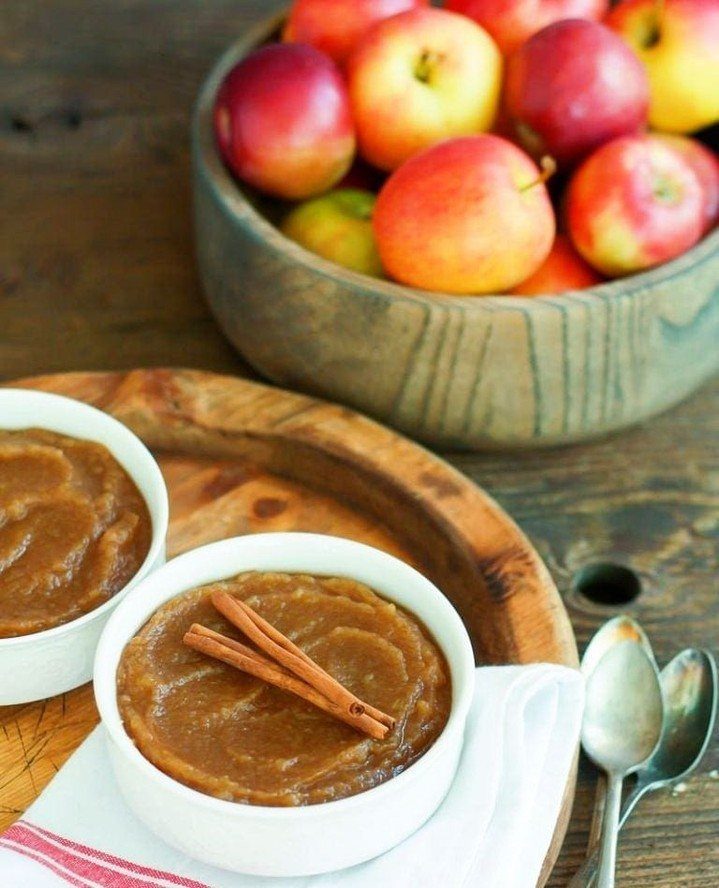 Brown cinnamon applesauce in white bowls with a bowl of fresh apples behind.