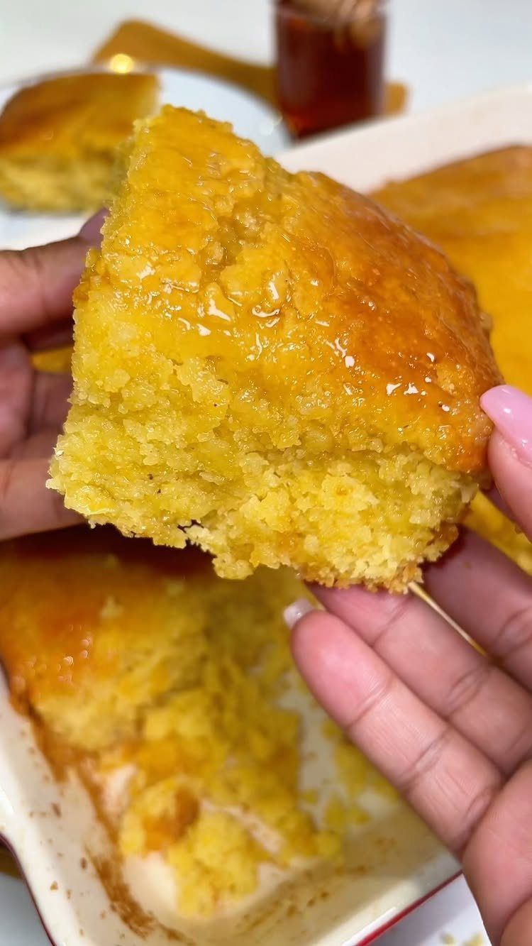 Golden-yellow glazed cornbread piece held by hands over a baking dish.