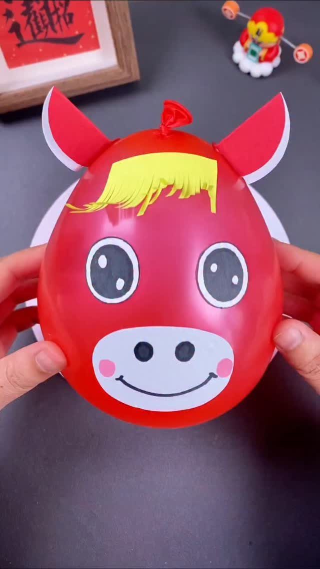 Red balloon horse craft with paper ears and yellow hair held by hands.